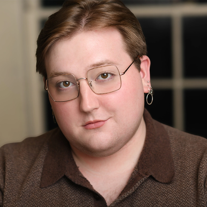 Margot May Waldron, a person with short light brown hair, wearing glasses and two earrings, poses indoors. They wear a brown collared shirt and have a neutral facial expression. The blurred background shows a window with black panes.