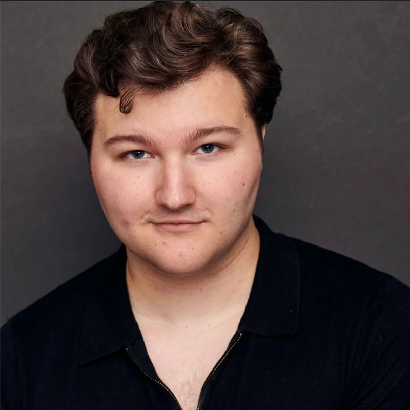 A young man with short, wavy brown hair and light skin looks at the camera with a slight smile. He is wearing a black collared shirt and is posed in front of a plain dark gray background, reminiscent of the granite hues of Stone Mountain, Georgia.