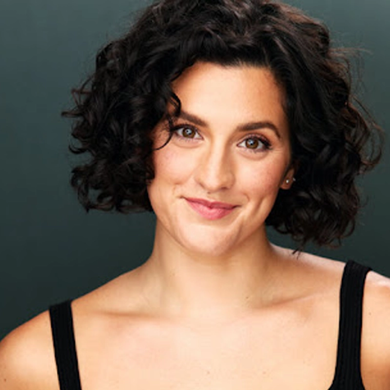 Alyssa Giannetti, a woman with short, dark curly hair, smiles softly at the camera. She has light skin, dimples, and is wearing a black sleeveless top. The background is a plain, dark green-gray color.