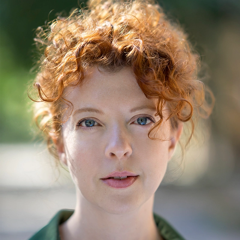 Bethany Geraghty, a woman with fair skin and curly red hair, gazes directly at the camera with a calm expression. She wears a green top and stands outdoors against a softly blurred green background that enhances her natural features.
