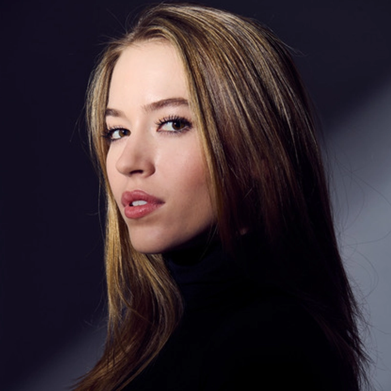 A young woman with long, straight, highlighted brown hair poses against a dark background, wearing a black turtleneck. She looks confidently at the camera with a slight smile and neutral makeup, illuminated by soft, angled lighting.