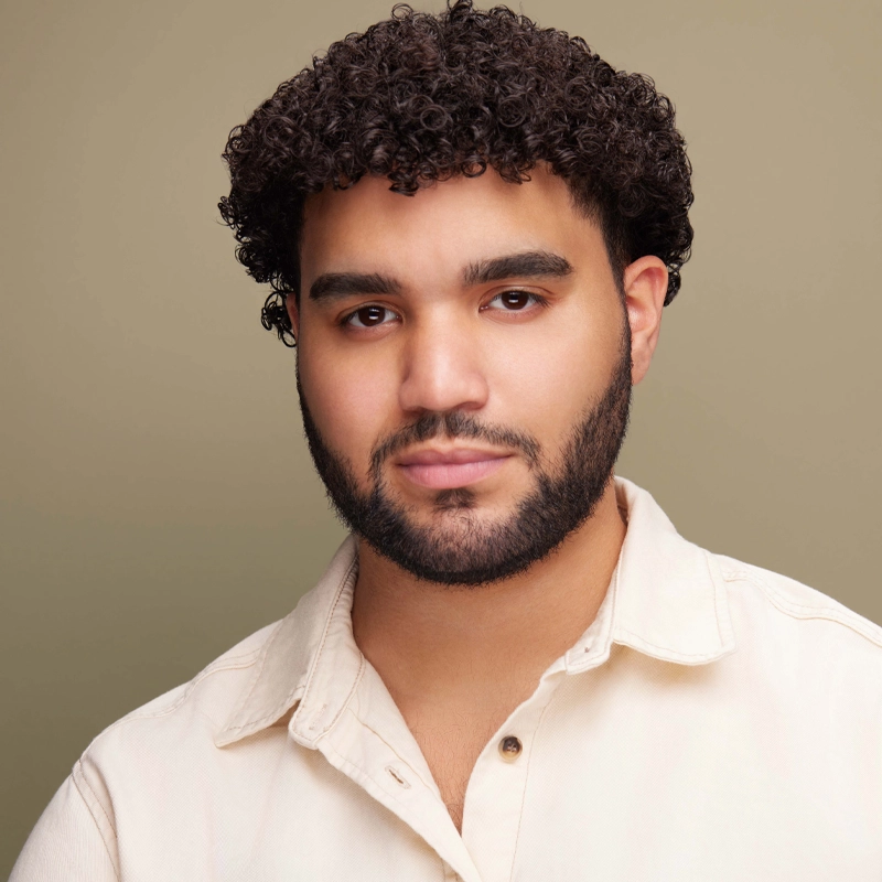 A young man with short, curly dark hair and a neatly trimmed beard looks at the camera with a neutral expression. He is wearing a cream-colored collared shirt against a plain beige background. The lighting is soft and even.