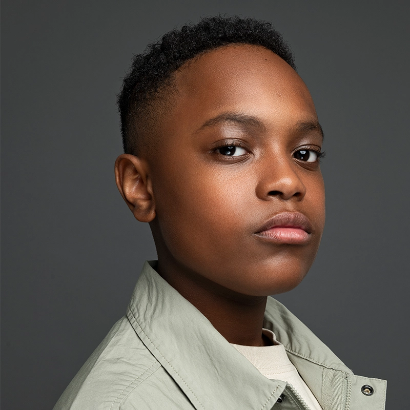 A young Black boy with short curly hair looks confidently at the camera. He wears a light greyish-green jacket over a beige shirt. The dark gray background highlights Davis Matthews’ calm and serious expression.