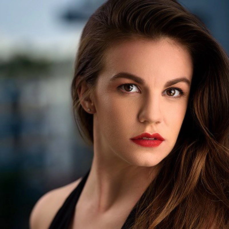 A woman with long brown hair, parted to the side, gazes confidently at the camera. Emma Grimsley wears a black top, red lipstick, and subtle makeup. The softly blurred background hints at an outdoor or urban setting.