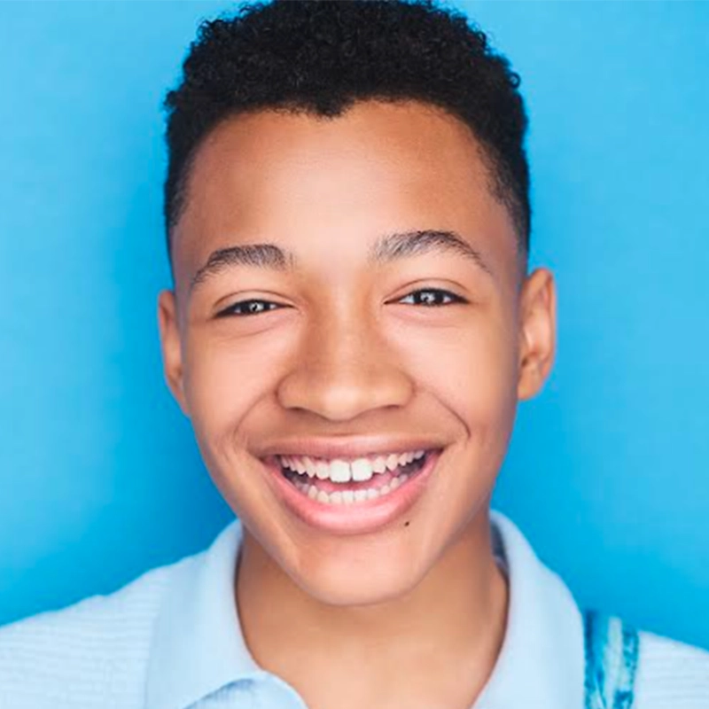 A smiling teenage boy with short curly hair stands in front of a bright blue background. Ethan Joseph is wearing a light blue collared shirt and looking directly at the camera, showing his teeth in a wide, cheerful smile.