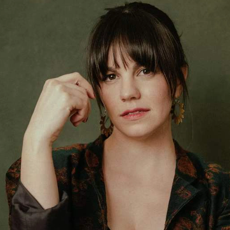 Emma Grimsley, with straight dark hair and bangs, looks at the camera. She wears large sun-shaped earrings and a patterned dark jacket, her hand near her face in a neutral pose against an olive-green background.