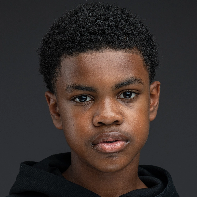 A young Black boy with short, curly hair looks directly at the camera. He has a calm, neutral expression and wears a black hoodie. The plain dark background and soft lighting highlight his features, reminiscent of portraits like those of Jackson Hayes.