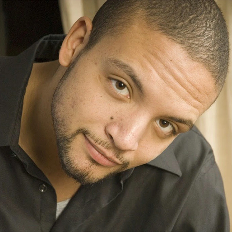 Joseph Reese Anderson, a man with short hair and a trimmed beard, looks at the camera, slightly tilting his head. He has light brown skin, a small mole on his right cheek, and wears a black collared shirt over a gray top against a softly blurred background.