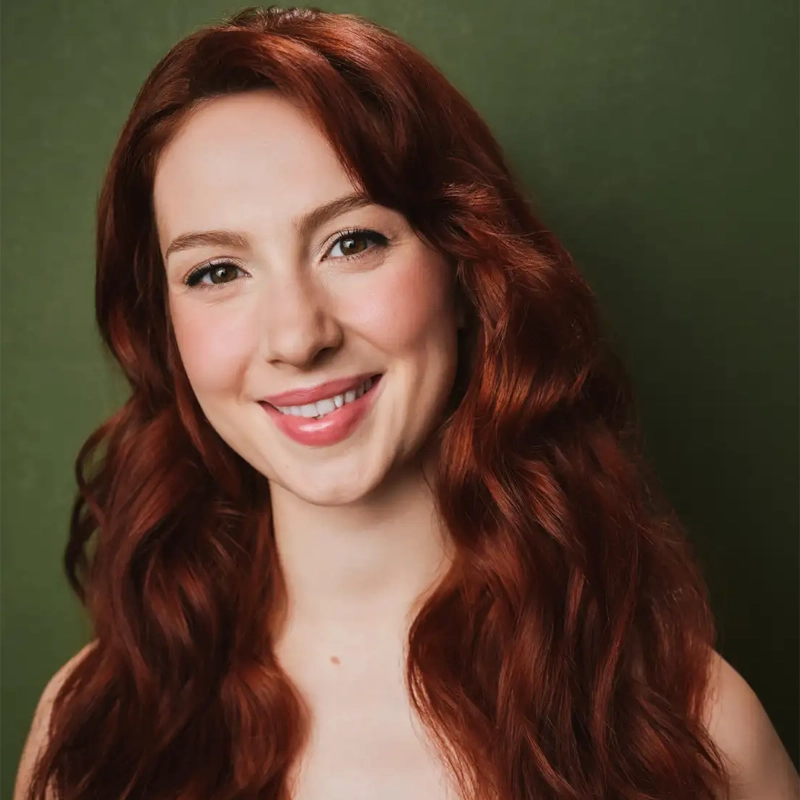 A young woman with long, wavy auburn hair smiles warmly at the camera. She has light skin and brown eyes, posed against a solid, muted green background.