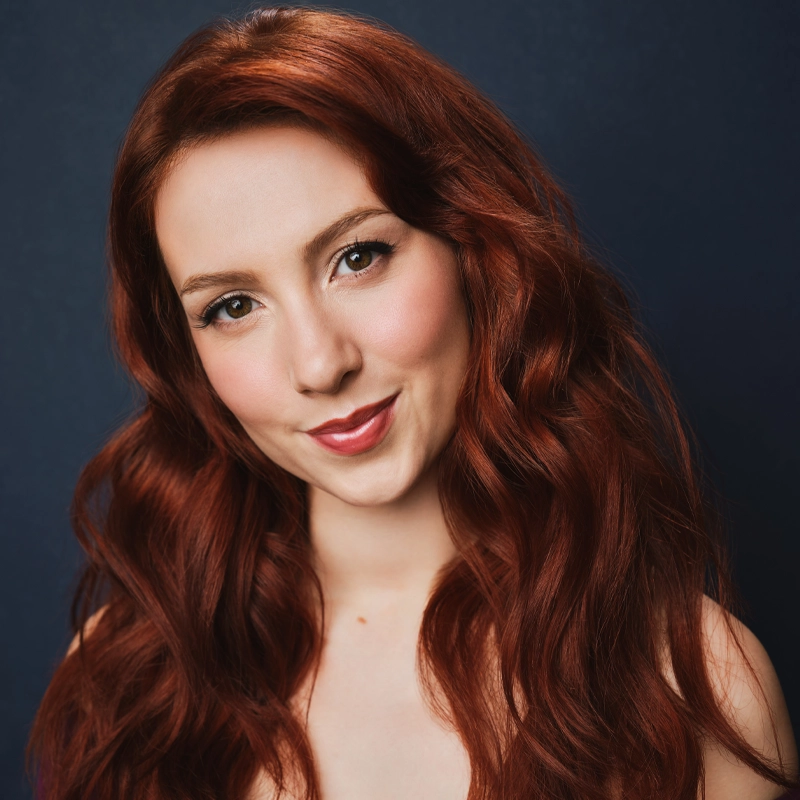 A woman with long, wavy auburn hair smiles gently. She has fair skin, rosy cheeks, and wears natural makeup. She faces the camera against a plain dark blue background, her head slightly tilted, exuding warmth and friendliness.