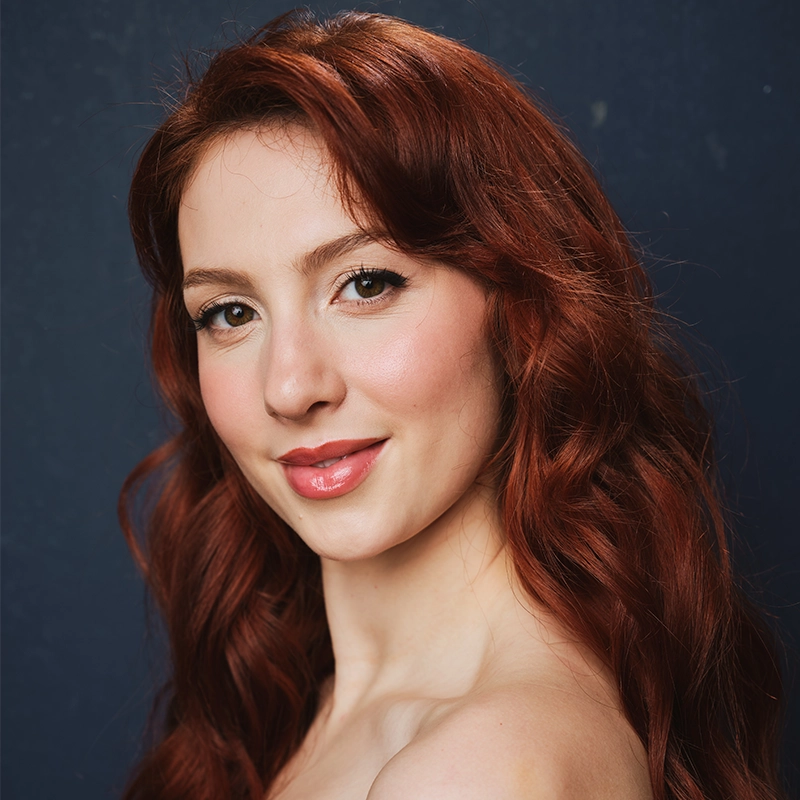 A young woman with long, wavy red hair poses against a dark blue background. She has fair skin, light makeup with pink lipstick, and a soft smile. Her head is slightly tilted, and her bare shoulders are visible.