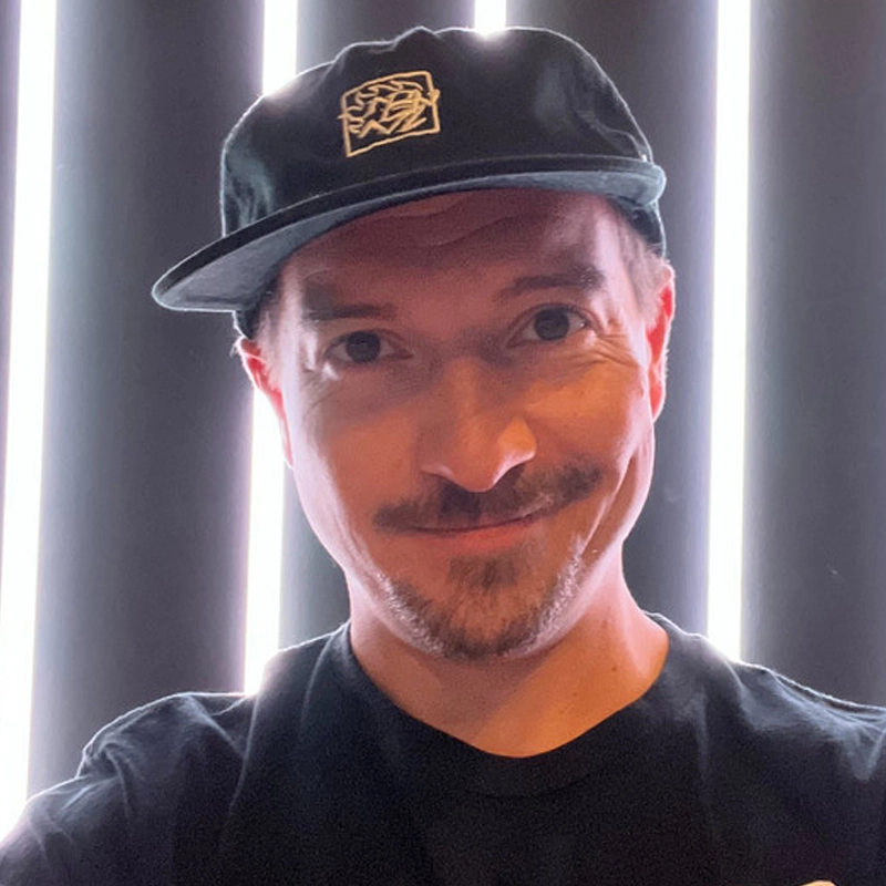 Michael Berberich, a man with light skin, a mustache, and a short beard, smiles at the camera. He wears a black cap with a white abstract logo and a black T-shirt. Vertical light beams shine behind him, creating a bright, modern background.