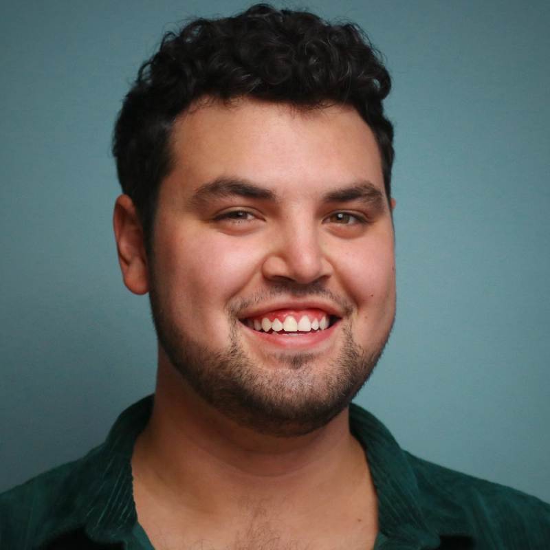 A young man with curly dark hair and facial hair smiles warmly at the camera. He is wearing a green collared shirt and stands in front of a plain teal background. The lighting is soft, highlighting his friendly expression.