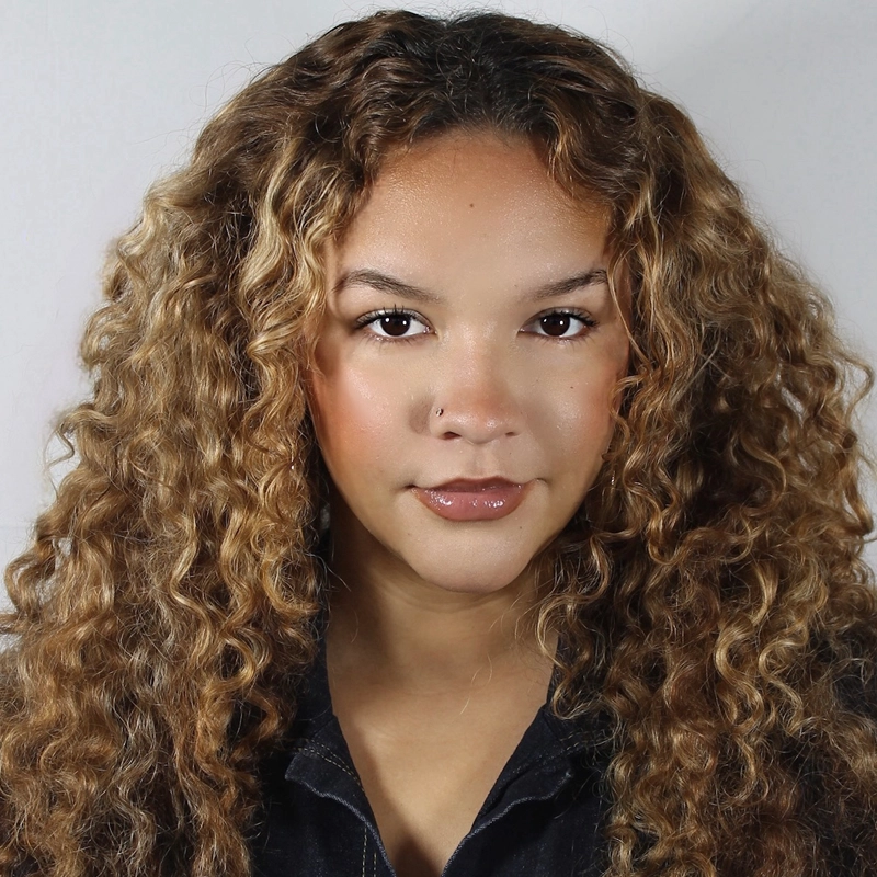 A person with long, voluminous, curly light brown hair looks directly at the camera. They have warm-toned makeup, glossy lips, and a small nose ring. They are wearing a dark top and are posed against a plain light-colored background.