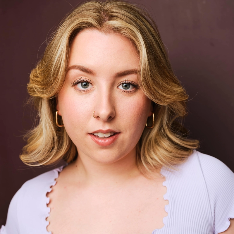 A young woman with wavy blonde hair and blue eyes looks at the camera. Ryanne Solinsky wears gold hoop earrings, a light purple, scallop-edged top, and a small nose ring. The plain dark background enhances her brightly lit face.
