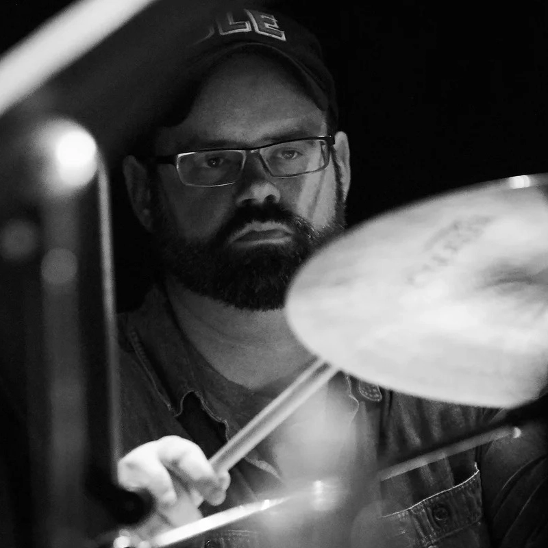 A bearded man wearing glasses and a cap plays drums. He holds drumsticks and looks focused. The image is black and white, with blurred drum equipment in the foreground and a dark background.