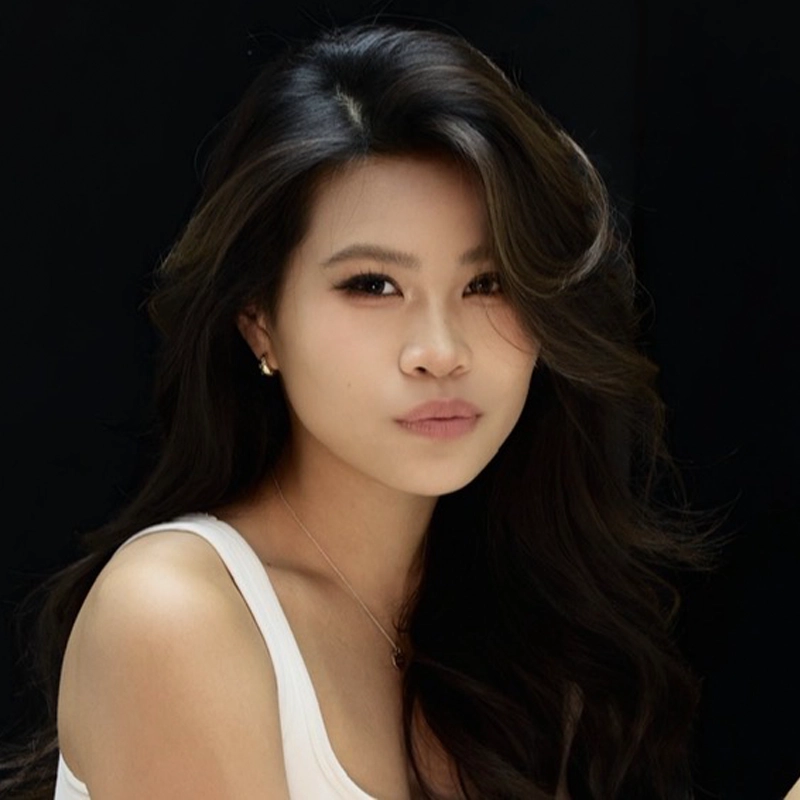 Alyssa Wong, a woman with long, wavy dark hair, poses against a black background. She wears a white sleeveless top and small earrings. Her expression is neutral, and she looks directly at the camera with soft, natural makeup.