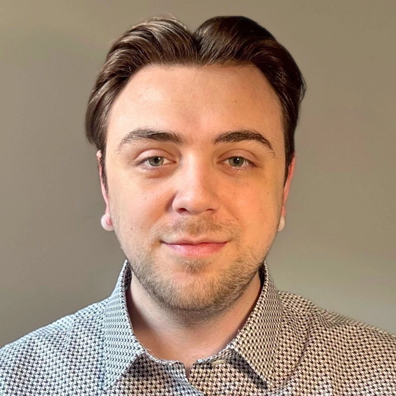 A young man with fair skin, short brown hair, and blue eyes is facing the camera and smiling slightly. Caleb Conaway is wearing a light gray patterned button-up shirt. The background is plain and softly lit in neutral beige tones.