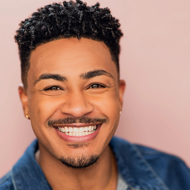 Morgan Perry, a man with short, curly black hair and a trimmed goatee, smiles broadly, showing his teeth. He wears a denim jacket over a gray shirt and small hoop earrings. The soft pink background and bright lighting highlight his cheerful expression.