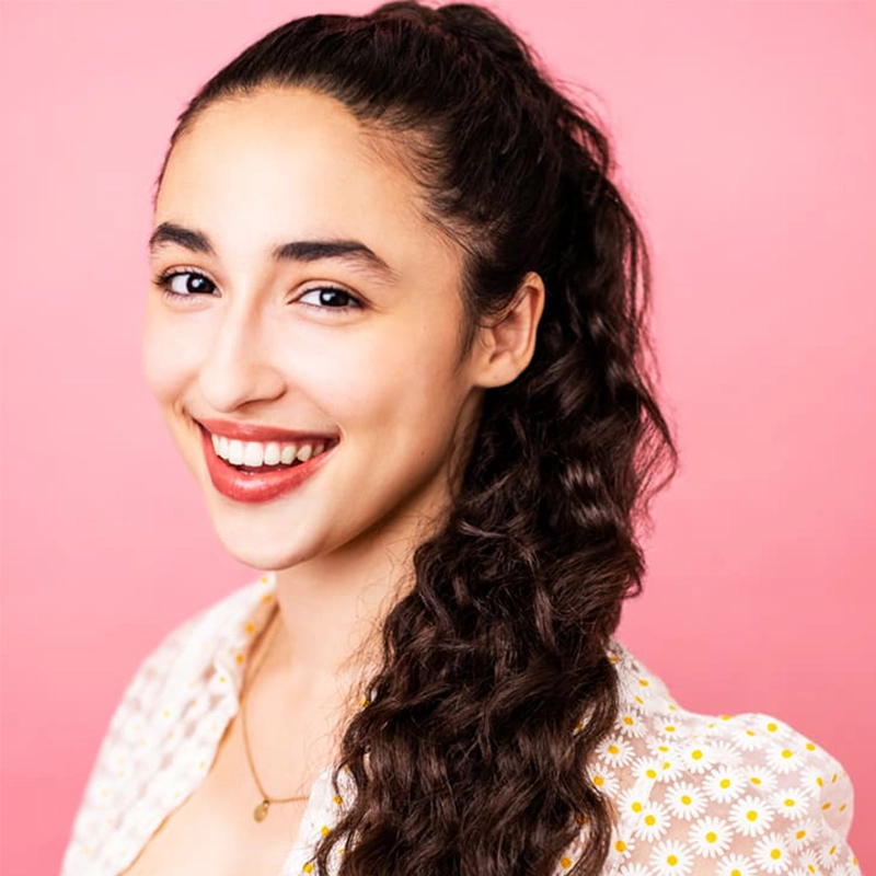 A young woman with long, curly brown hair in a high ponytail smiles at the camera. She wears a white blouse with yellow daisy prints and a gold necklace. The background is bright pink, creating a cheerful, vibrant atmosphere.