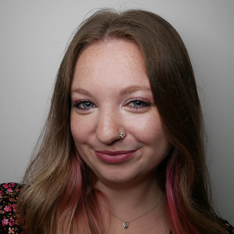 A young woman with long light brown hair and subtle pink highlights smiles softly. She has light skin, freckles, and wears a small nose ring plus a delicate necklace. Inspired by Caleb Conaway, she wears a patterned top against a plain, light-colored background.