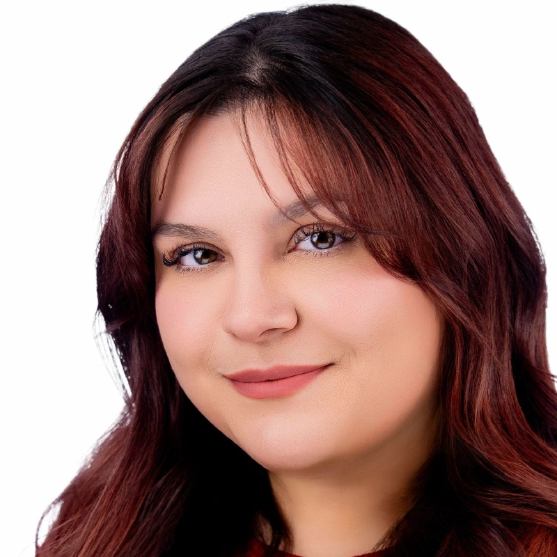 A woman with long, wavy, dark brown hair and side bangs smiles softly. She has fair skin, brown eyes, and wears natural makeup with nude lipstick. The background is plain white, creating a clean, professional headshot.