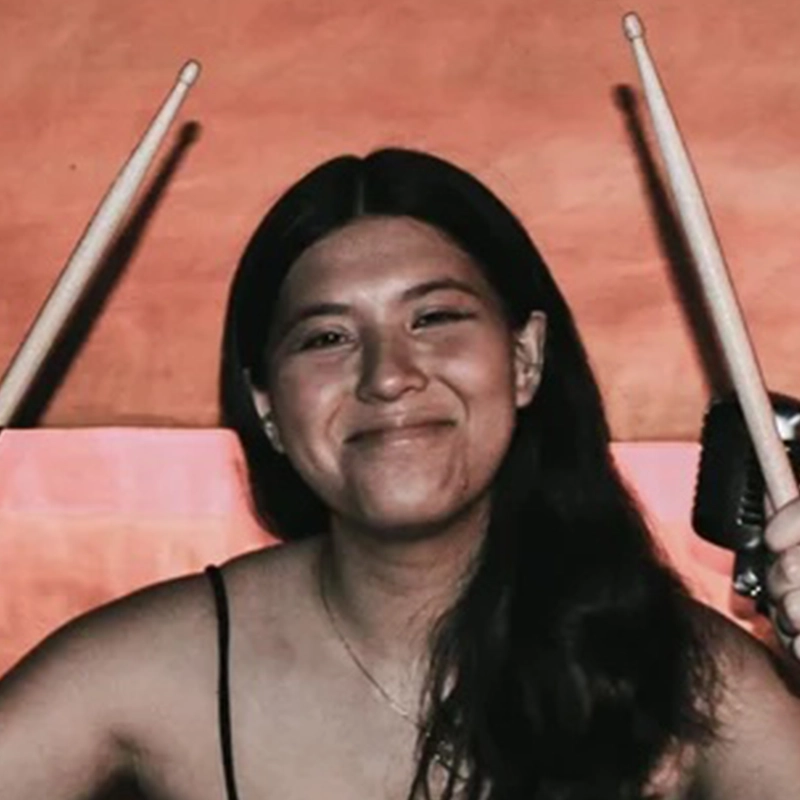 A young woman with long dark hair smiles at the camera, holding drumsticks in both hands raised in the air. She wears a sleeveless top and stands against a warm orange and pink background, channeling Caleb Conaway’s vibrant stage energy. A microphone is partially visible to her right.