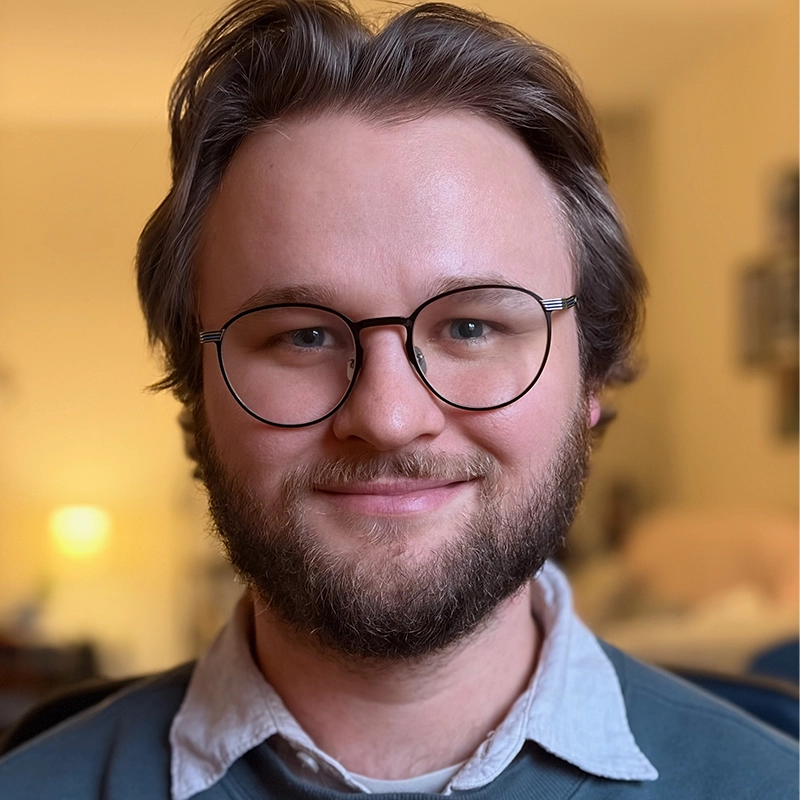 Caleb Conaway, a young man with light skin, short brown hair, a beard, and round glasses, smiles at the camera. He wears a blue sweater over a collared shirt in a cozy, warmly lit indoor setting with softly blurred furnishings.