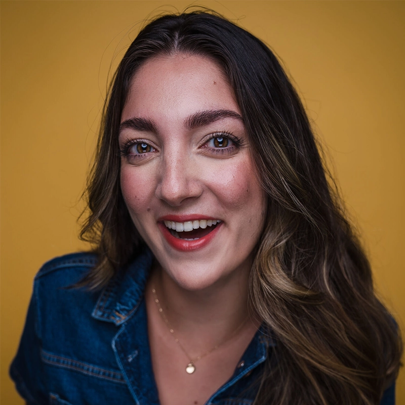 A young woman with long, wavy brown hair smiles at the camera. She wears a denim shirt and a delicate gold necklace. The background is a solid mustard yellow, giving a warm and cheerful vibe to the portrait.