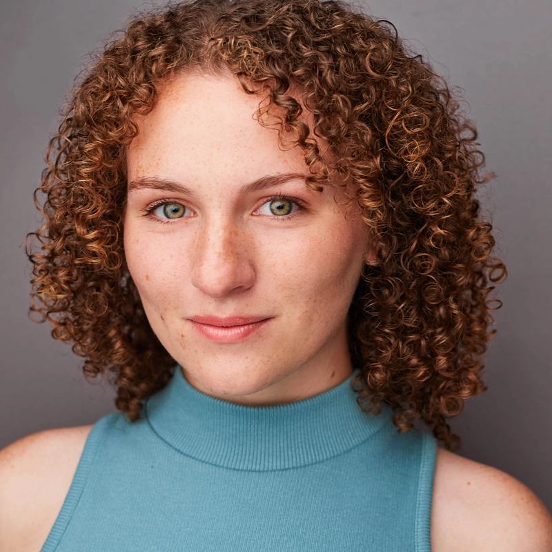 Megan Collina, a young woman with fair skin and curly, light brown hair, looks at the camera in a sleeveless, high-neck teal top. She stands against a plain gray background, her expression calm and confident.