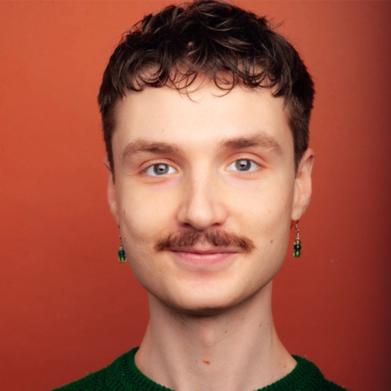 Cullen Curth, a young person with short brown hair, blue eyes, and a brown mustache, smiles at the camera. They wear a green top and small dangling earrings with green beads against a solid reddish-orange background.