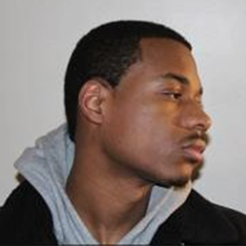 A young man with short black hair and a trimmed mustache and beard looks to his right. He is wearing a gray hoodie layered under a black jacket. The background is plain and light-colored. The image is a close-up of his face and upper shoulders.