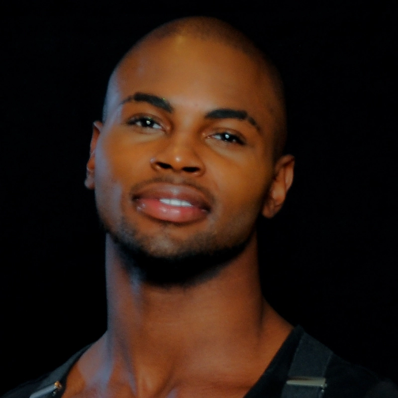 Morgan Perry, a young man with smooth dark skin and a shaved head, faces the camera, smiling gently with closed lips. He has well-groomed eyebrows, trimmed facial hair, and wears a dark off-shoulder shirt against a black background.