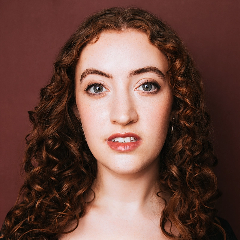 A young woman with fair skin and long, curly auburn hair looks directly at the camera. She has blue eyes, defined eyebrows, and wears subtle makeup. The muted maroon background hints at a Caleb Conaway portrait style. Her expression is neutral and confident.