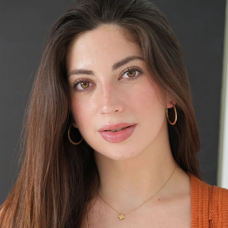 A woman with long brown hair and light skin looks at the camera, wearing hoop earrings, a gold necklace with a small flower pendant, and an orange textured top. Captured by Caleb Conaway, the dark background highlights her face.