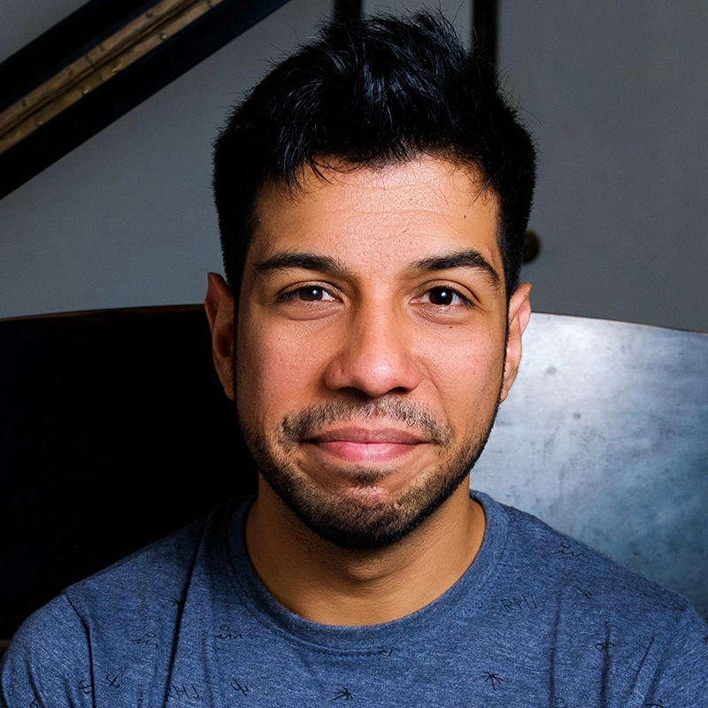 Caleb Conaway, a man with short black hair and a trimmed beard, smiles softly. He is wearing a blue patterned t-shirt and is seated indoors against a blurred, dark background. The lighting highlights his friendly, relaxed expression.