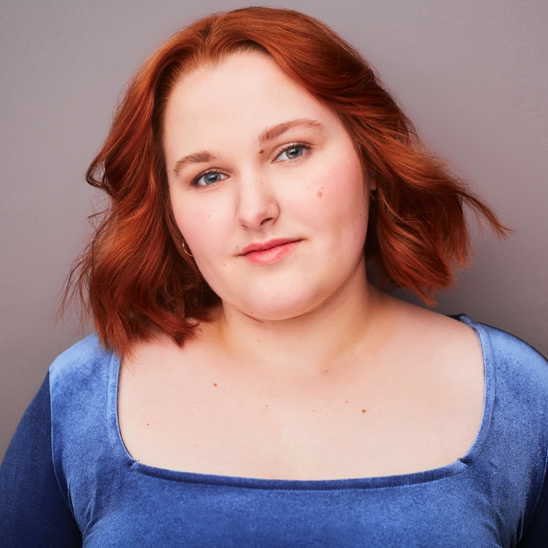 Amelia Firestone, with wavy, shoulder-length red hair and fair skin, smiles softly at the camera. She wears a light blue velvet top with a square neckline, standing confidently against a plain gray backdrop.