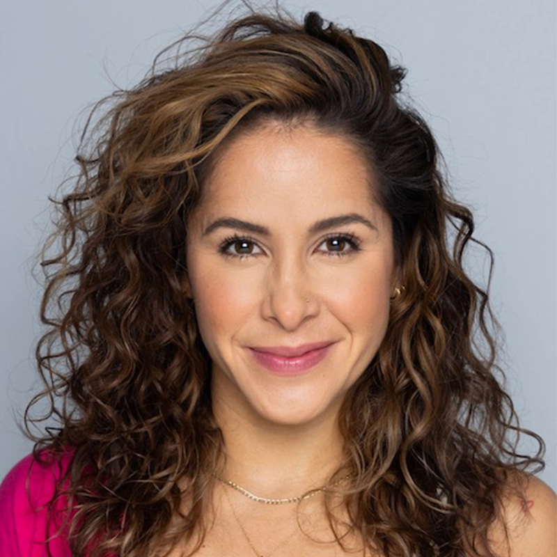 A woman with medium skin and long, curly brown hair smiles at the camera. She wears a pink top, subtle makeup, and two delicate gold necklaces. The background is a neutral light gray. Her expression is friendly and inviting.