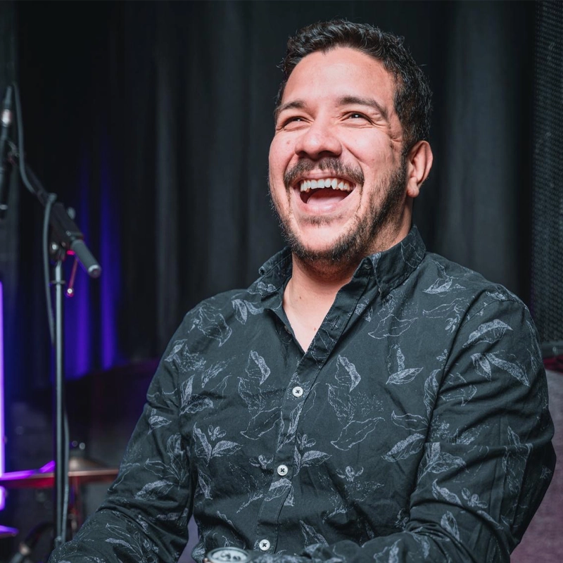 Daniel Gutierrez, a man with short dark hair and a trimmed beard, is smiling widely and laughing. He wears a dark patterned button-up shirt. The dimly lit purple background and microphone stand create a joyful, relaxed atmosphere.