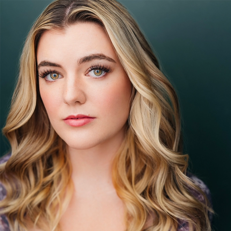 A young woman with long, wavy blonde hair and fair skin looks directly at the camera. She has green eyes, subtle makeup with pink lipstick, and wears a purple patterned top. The background is a smooth gradient of dark green.