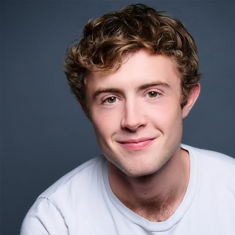 A young man with wavy light brown hair and hazel eyes smiles gently. He wears a plain white t-shirt and sits against a plain dark gray background. The lighting is soft, highlighting his facial features and creating a friendly, approachable appearance.