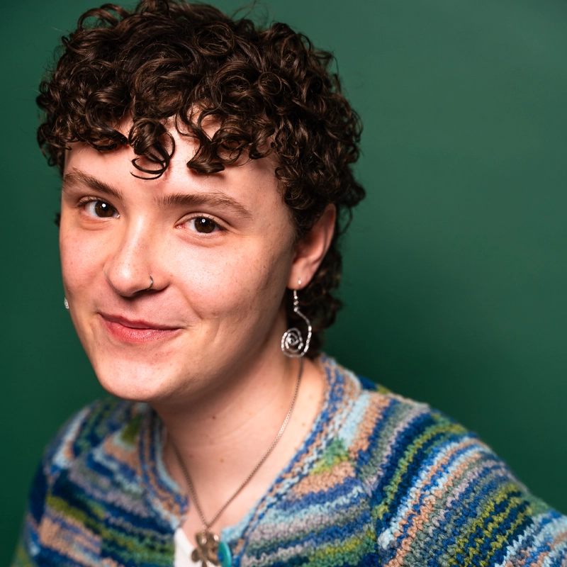 Walter Higginbotham, with short, curly brown hair, smiles gently at the camera. He wears a blue, green, and white striped sweater, a spiral earring, a necklace, and nose ring against a solid teal green background.