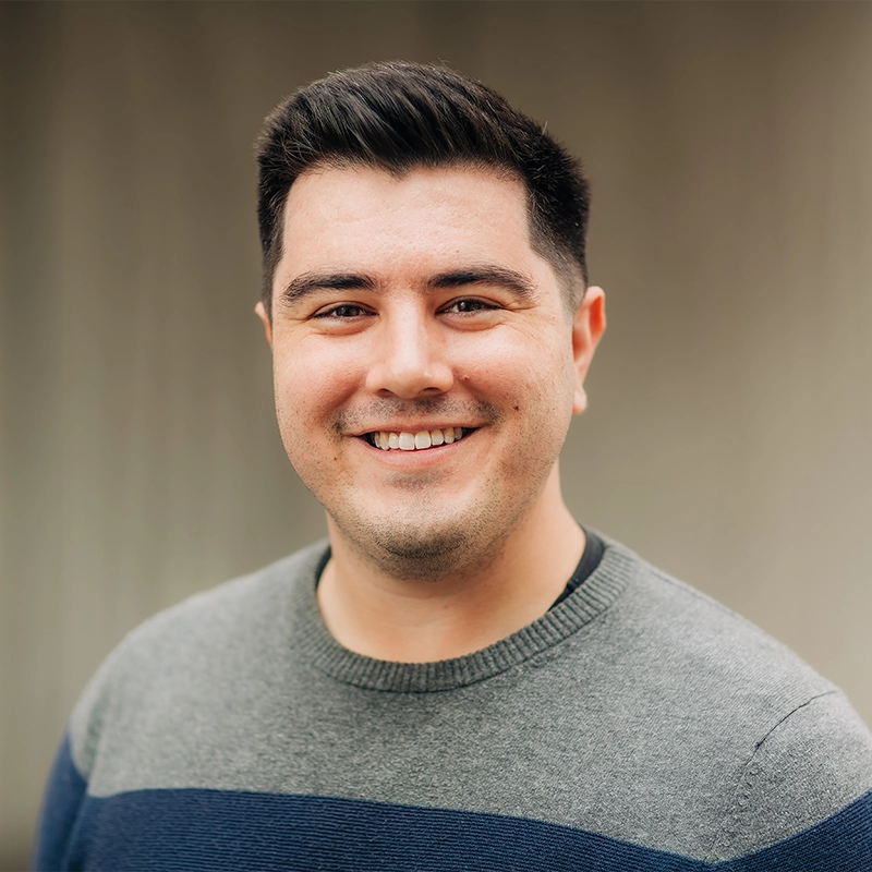 Jason Belanger, a young man with short dark hair and a trimmed beard, smiles at the camera. He is wearing a gray sweater with a navy blue stripe, while the blurred background draws focus to his friendly expression.