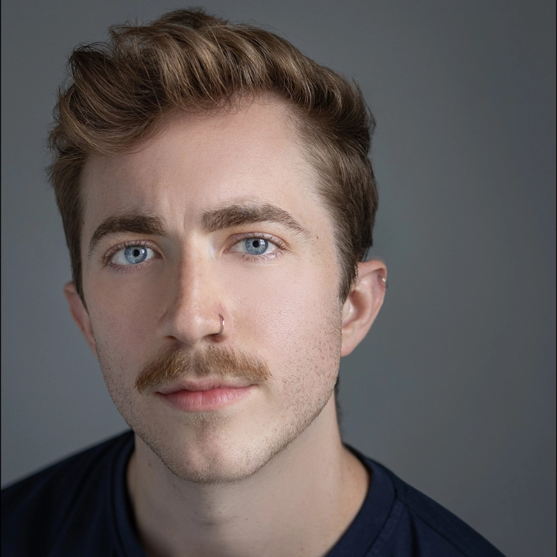 A young man with light brown hair, blue eyes, and a light mustache looks directly at the camera. He has a nose ring in his left nostril and wears a dark-colored shirt. The background is plain and gray, with soft lighting on his face.