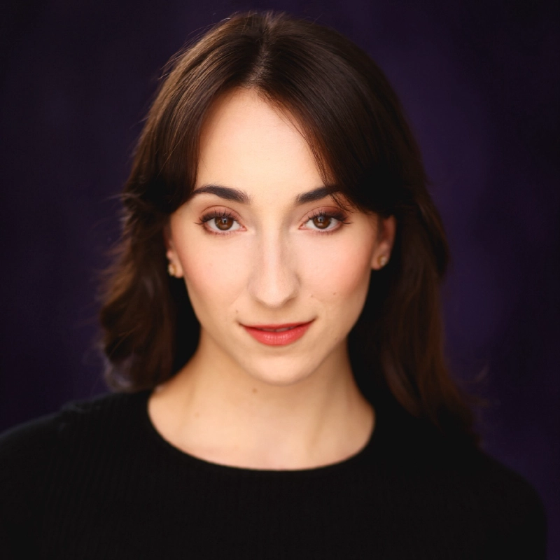 A woman with fair skin, long brown hair, and brown eyes looks directly at the camera. She wears a black top, subtle makeup with red lipstick, and small earrings. The background is a deep purple gradient, and the lighting is soft and even.