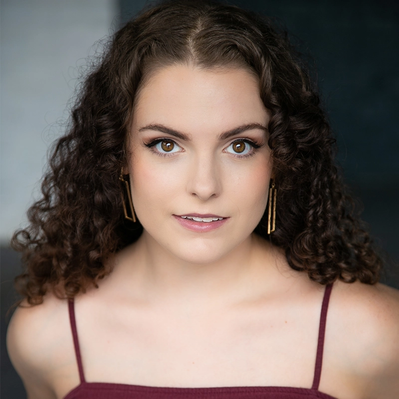 A young woman with fair skin and brown, curly hair looks directly at the camera. She wears gold rectangular earrings and a sleeveless, burgundy top with thin straps. The background is softly blurred in shades of dark blue and white.