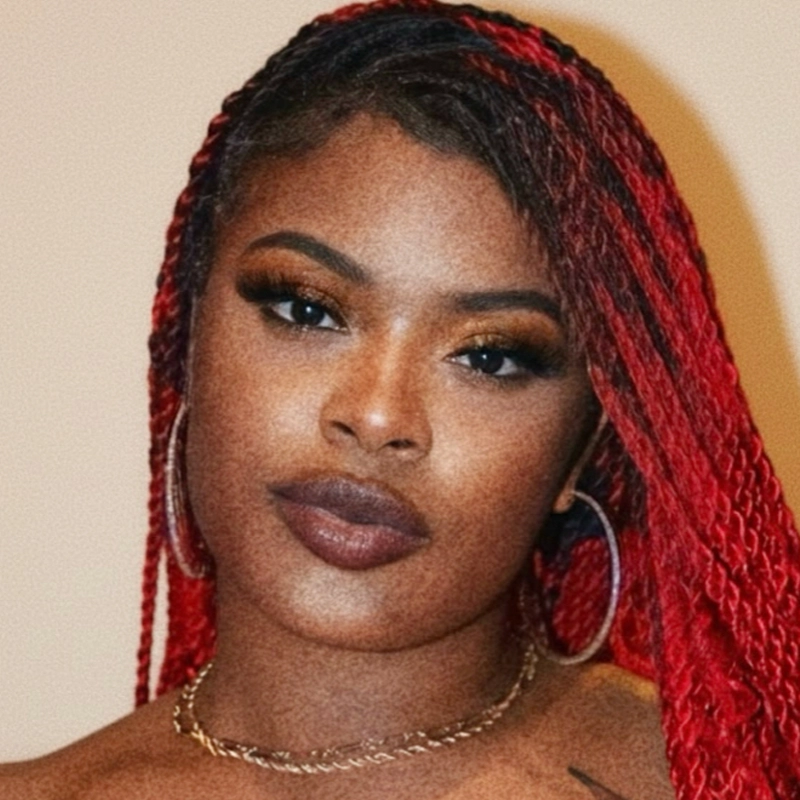 A woman with long, red braided hair looks at the camera. She has full eyebrows, defined eye makeup, long eyelashes, and dark lipstick. Peyton Leonard wears large hoop earrings and a thin gold chain necklace against a plain, light-colored background.