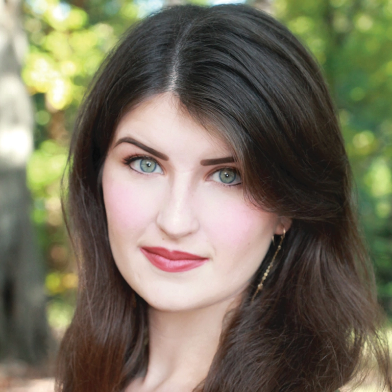 A young woman with fair skin, long brown hair, and green eyes looks at the camera. She has defined eyebrows, pink cheeks, and red lipstick. The background is blurred greenery, suggesting she is outdoors in a wooded area during daylight.