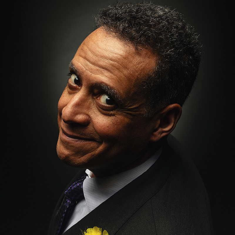 A middle-aged man in a dark suit and tie smiles warmly at the camera. He has short, curly hair and expressive eyes. A yellow flower is pinned to his lapel. The background is dark, highlighting Noel MacNeal’s face and friendly expression.