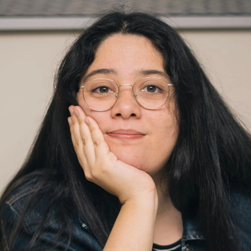 A person with long dark hair and round glasses rests their face on their hand, looking directly at the camera with a gentle smile. They wear a denim jacket and a striped top, evoking the laid-back style of Caleb Conaway. The background is neutral and out of focus.